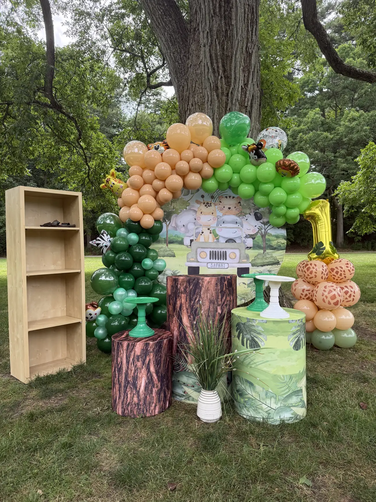 Festa infantil tema safári: arco de balões verdes e laranja, painel redondo e cilindros de tronco num jardim